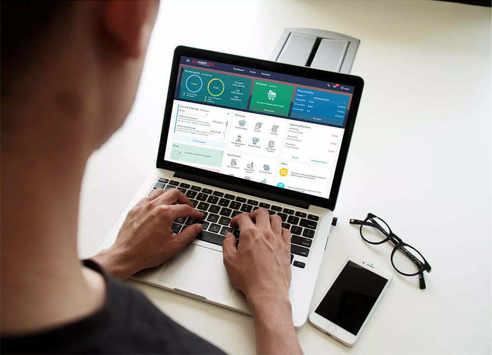A trader at a laptop showing the MintCFD account dashboard, phone and glasses on the desk